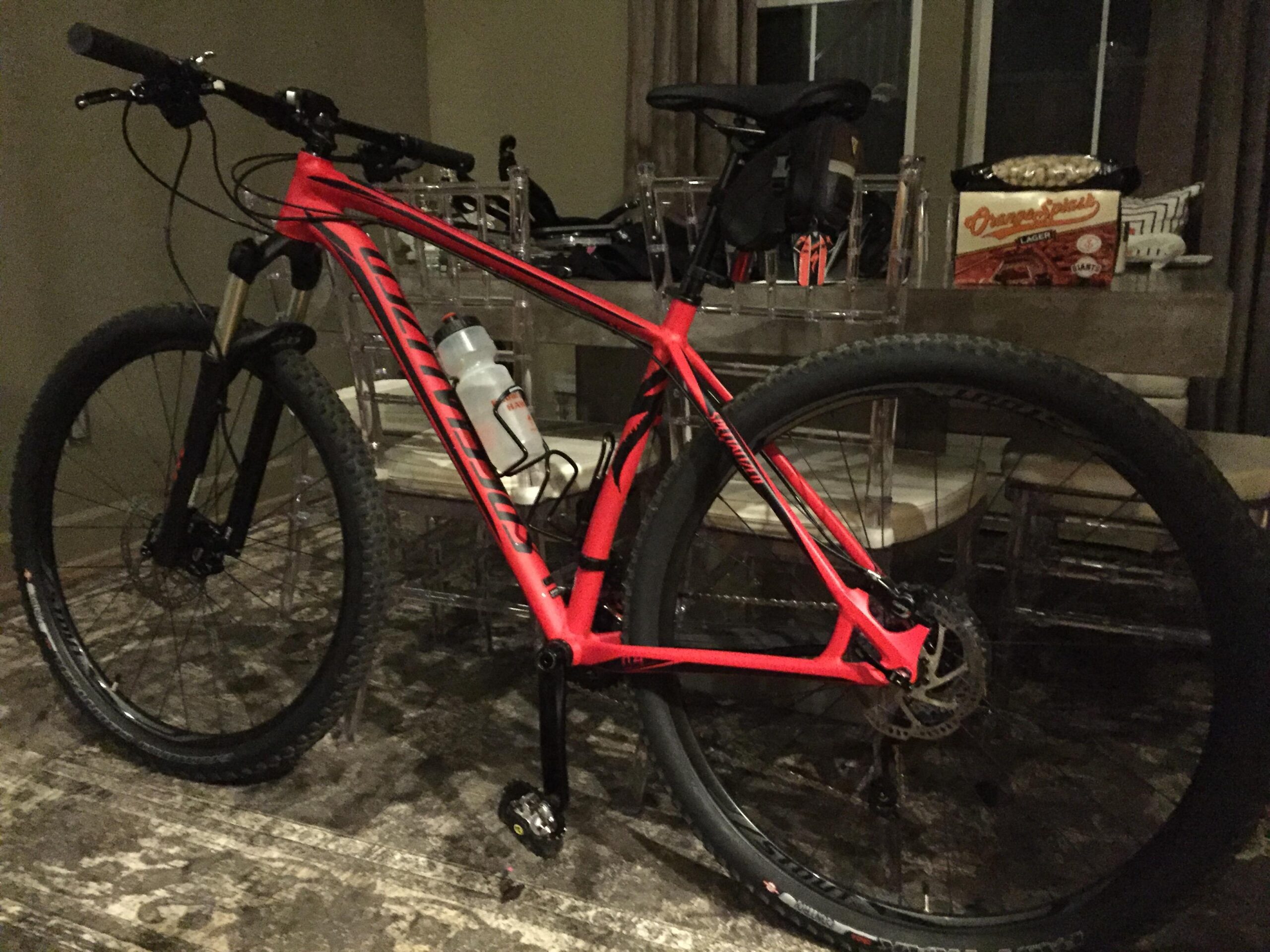 Specialized CRAVE 29: A close-up image of a bright red mountain bike with thick tires, positioned indoors. The bike features a water bottle attached to the frame. In the background, there is a table with a box of snacks and a few chairs. Soft lighting creates a cozy atmosphere.