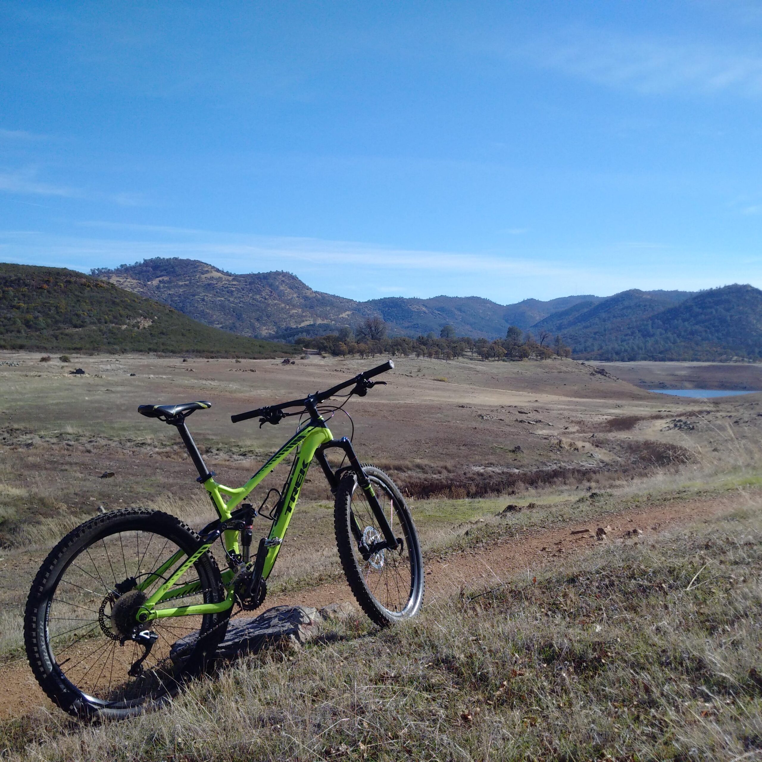 Trek Remedy 7 27.5: A bright green mountain bike is parked on a rocky patch near a dirt trail, with a vast landscape of dry grass and a distant lake. Rolling hills and mountains are visible in the background under a clear blue sky. The scene conveys a sense of adventure in nature.