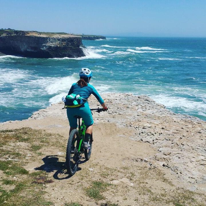A person in turquoise biking gear rides a green mountain bike along a rocky path at the edge of a cliff, overlooking the ocean. The waves crash against the shore, and the sky is clear and blue, indicating a sunny day. Wilder Ranch State Park mountain bike trail.
