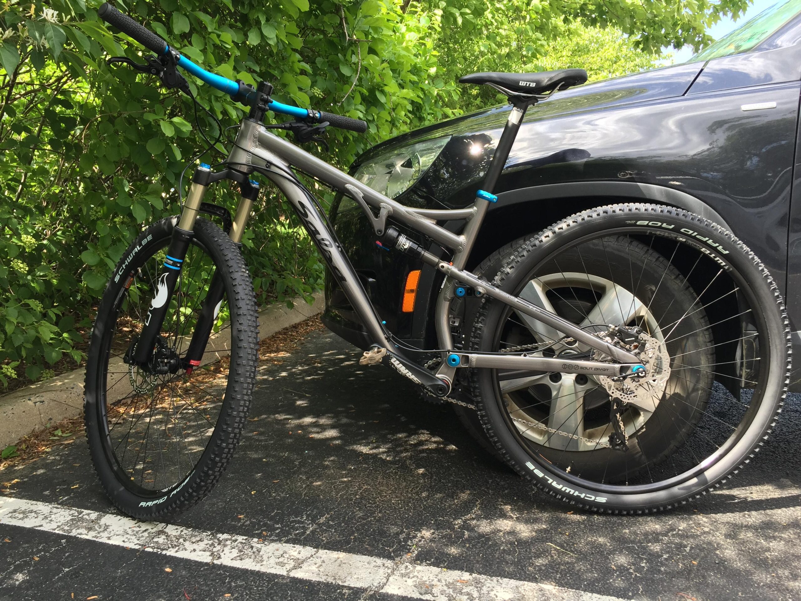 Salsa Spearfish: A mountain bike with a gray frame and blue accents is parked beside a black vehicle. The bike features thick tires with a prominent tread pattern, front suspension, and disc brakes. The background includes lush green foliage, indicating a natural outdoor setting.