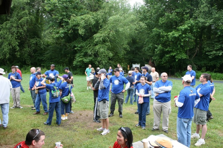 A group of diverse volunteers wearing matching blue t-shirts gathers in a grassy area surrounded by trees. Some individuals are conversing, while others are engaged with their phones or enjoying refreshments. The scene captures a sense of community and teamwork during an outdoor event.