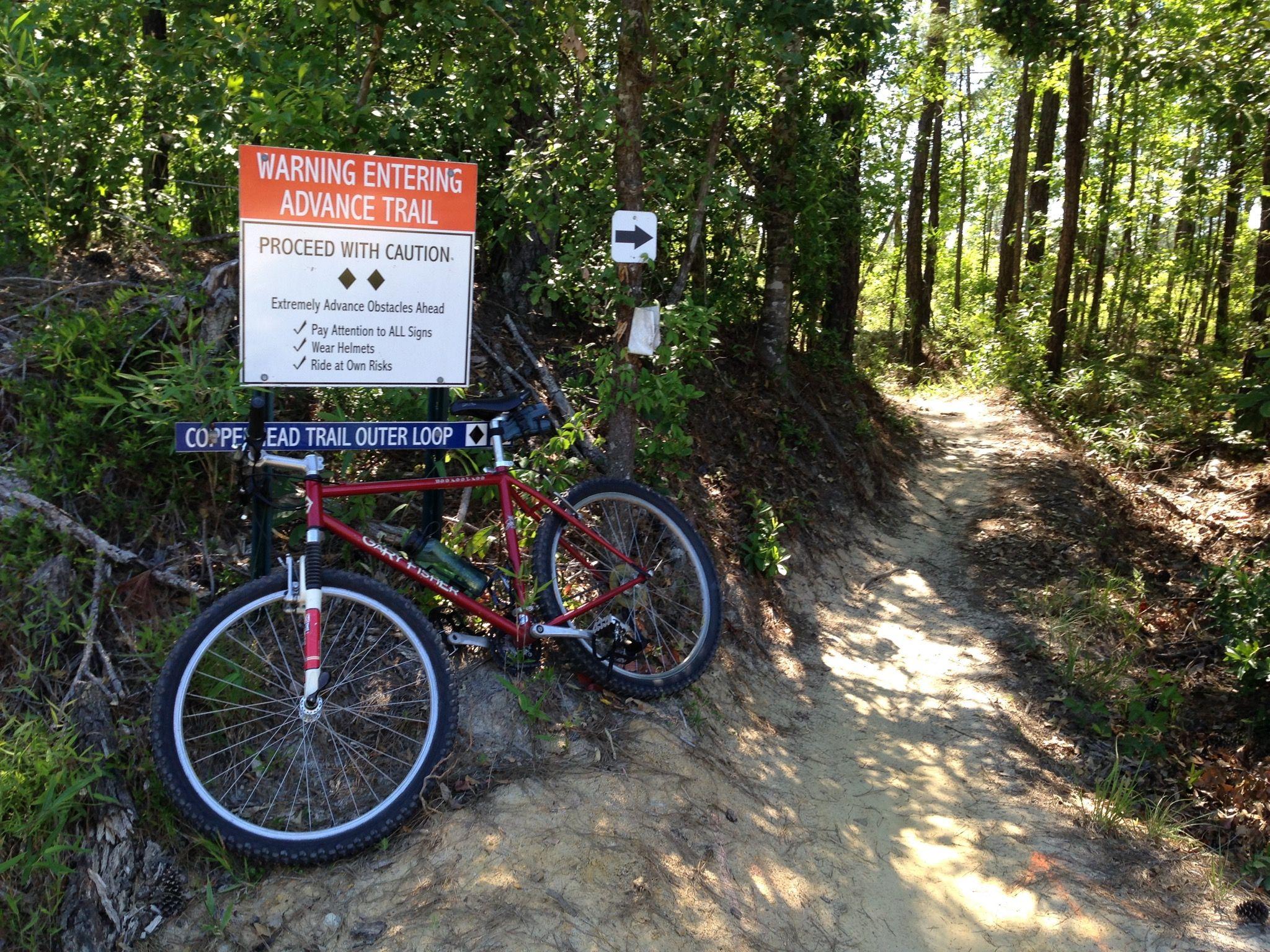 Gary Fisher Hoo Koo E Koo: A red mountain bike is parked next to a warning sign indicating the entrance to an advanced trail. The sign advises caution, mentions obstacles ahead, and emphasizes the importance of wearing helmets. The background features a wooded area with a narrow, sandy path leading further into the forest.