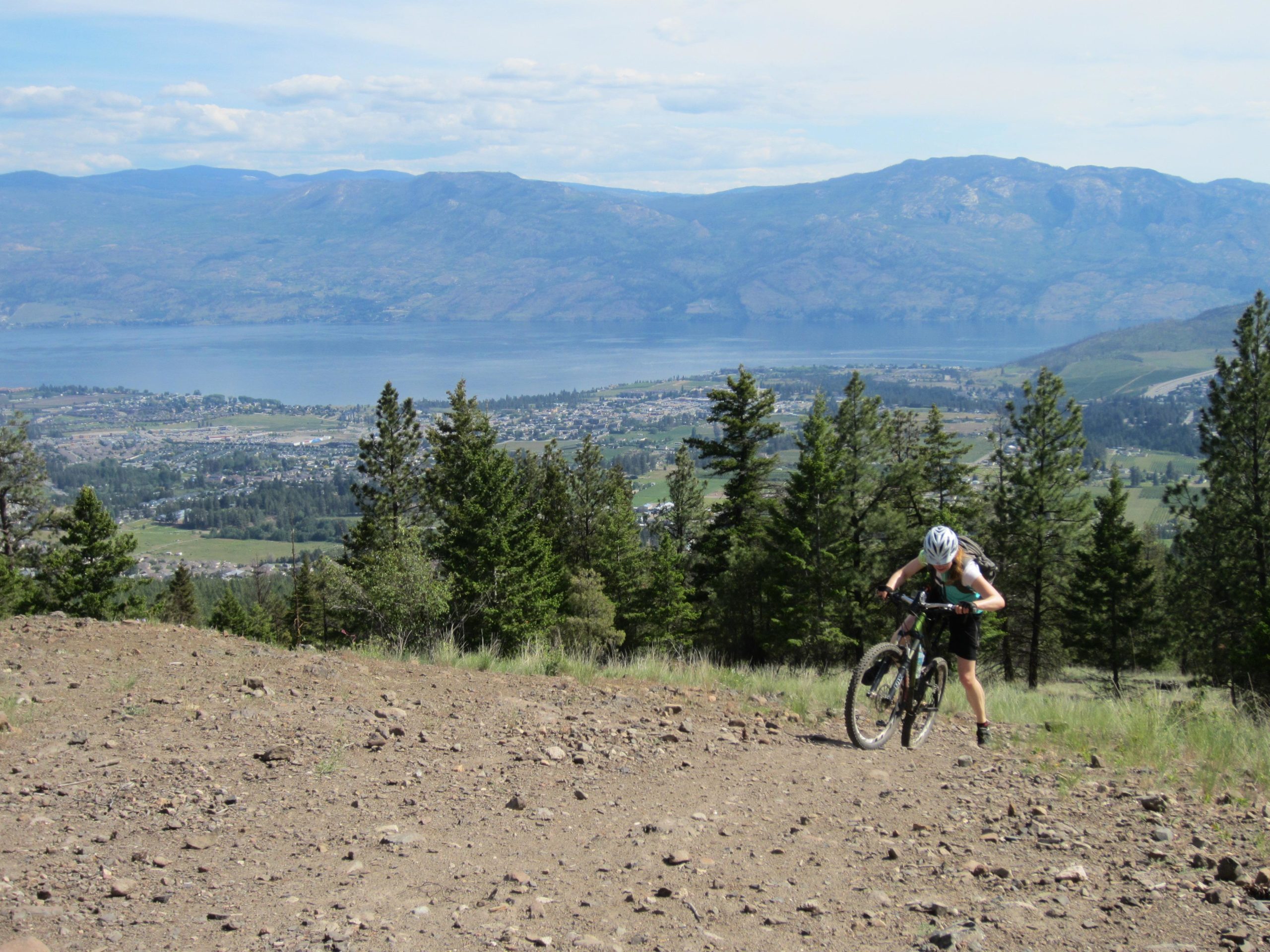A mountain biker struggles up a rocky trail in a forested area, with a panoramic view of a lake and mountains in the background on a sunny day. Smith Creek mountain bike trail.
