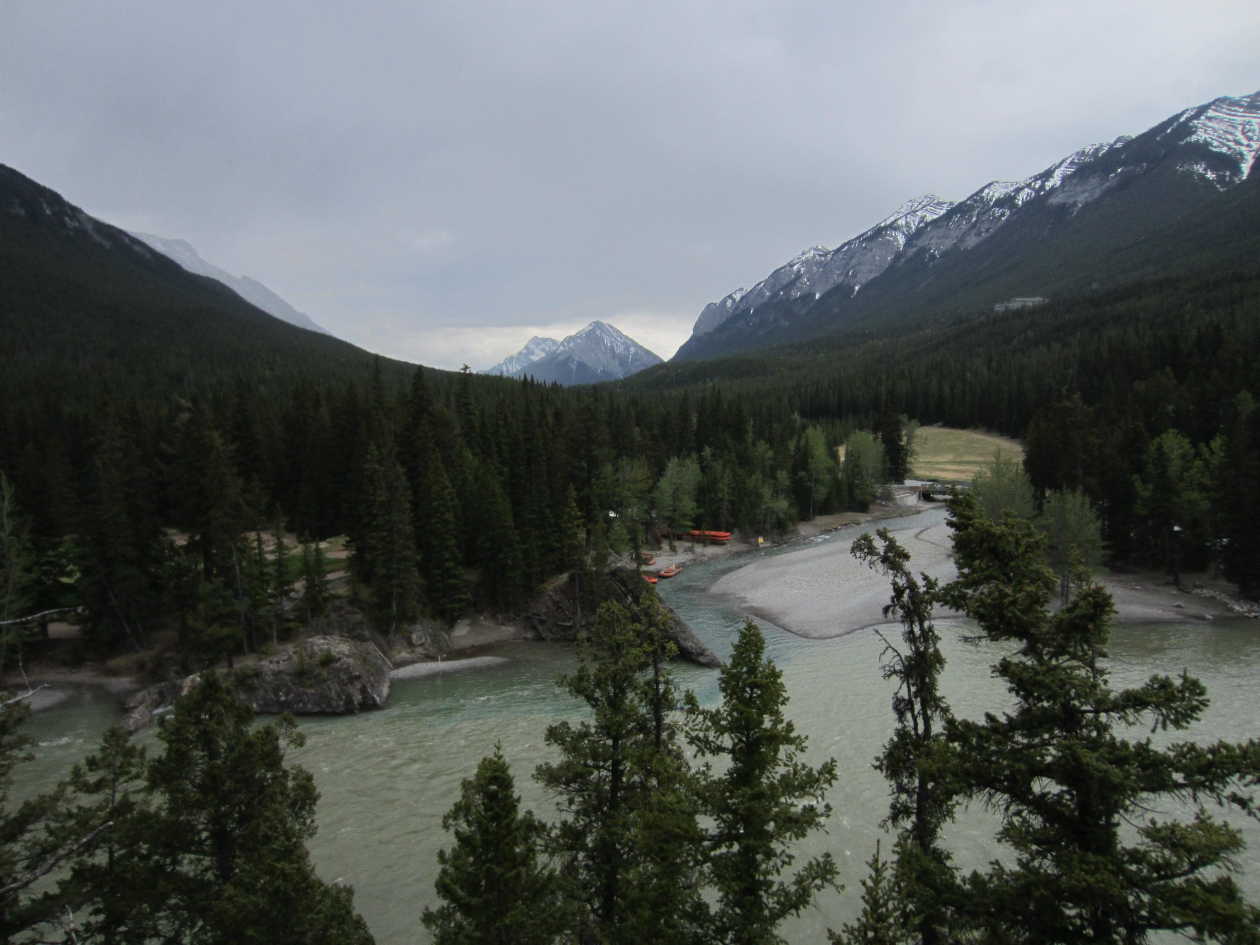 A scenic view of a river winding through a lush forest, surrounded by towering mountains. The landscape features green trees, a rocky shoreline, and some snow-capped peaks in the background under a cloudy sky. A small section of the riverbank shows a sandy area with a few canoes, suggesting a tranquil outdoor setting. Tunnel Mountain Trail System mountain bike trail.