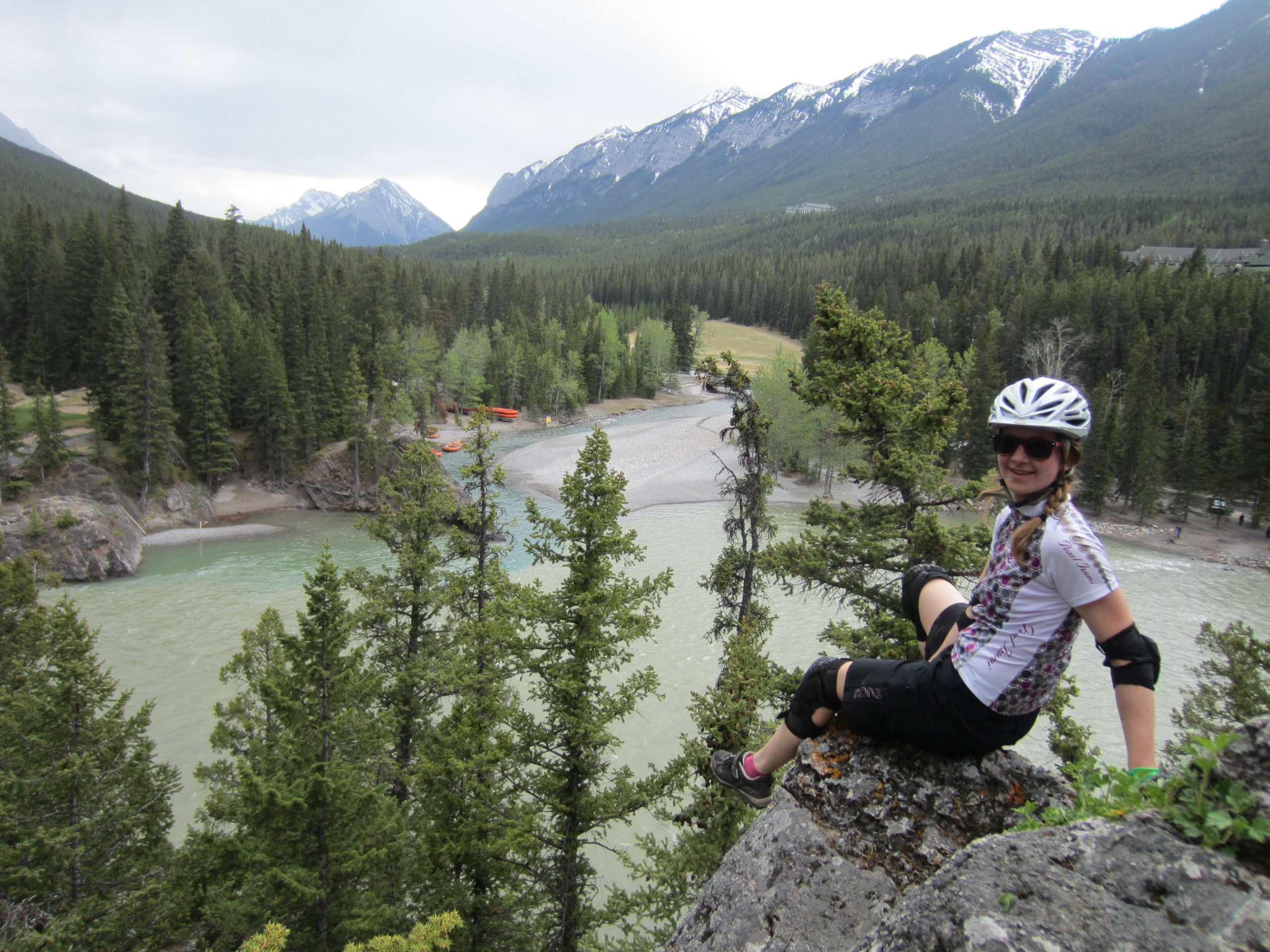 A person wearing a cycling helmet and protective gear sits on a rock overlooking a river surrounded by lush green trees and mountains. The scene shows a vibrant natural landscape with a mix of greenery and rocky terrain, and a glimpse of a beach area along the river. Tunnel Mountain Trail System mountain bike trail.