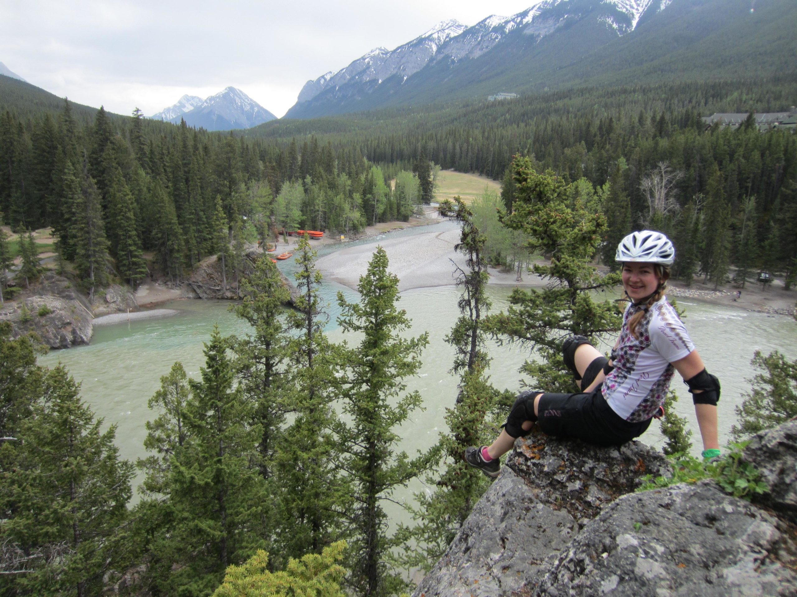A young girl in a cycling helmet and protective gear sits on a rocky ledge overlooking a river surrounded by trees and mountains. The scene captures a lush, green landscape with a hint of overcast sky, showcasing a serene outdoor environment. Tunnel Mountain Trail System mountain bike trail.