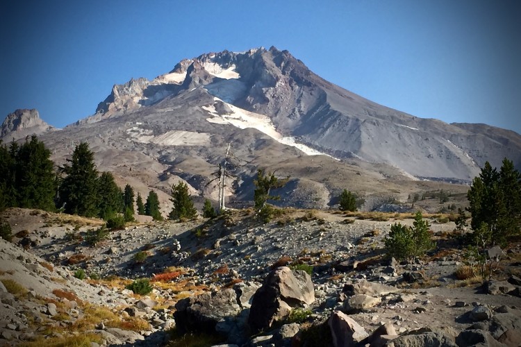 A view of a rugged mountain with a rocky landscape in the foreground, surrounded by pine trees. The sky is clear blue, and patches of snow are visible on the mountain's peaks, indicating a high altitude and possibly a volcanic origin. The terrain appears dry and rocky, with various shades of brown and green vegetation.