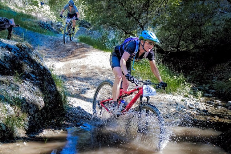 A mountain biker splashes through a puddle on a dirt trail, displaying excitement and determination. The rider, wearing a helmet and athletic gear, is in the foreground, while another cyclist can be seen in the background navigating the path. The lush surroundings include trees and rocks, suggesting a vibrant outdoor setting.