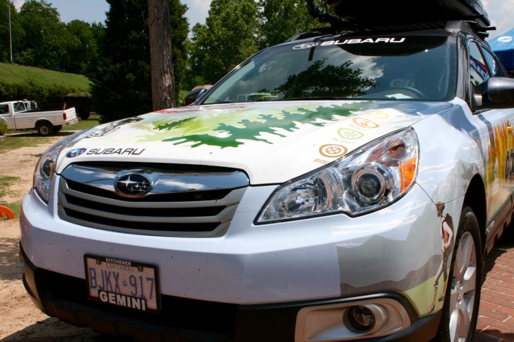 A Subaru vehicle with a nature-themed wrap featuring trees and outdoor icons, parked in a natural setting. The car's hood displays vibrant green graphics, and the Subaru logo is prominently visible on the front. A clear blue sky and trees are in the background, indicating an outdoor atmosphere.