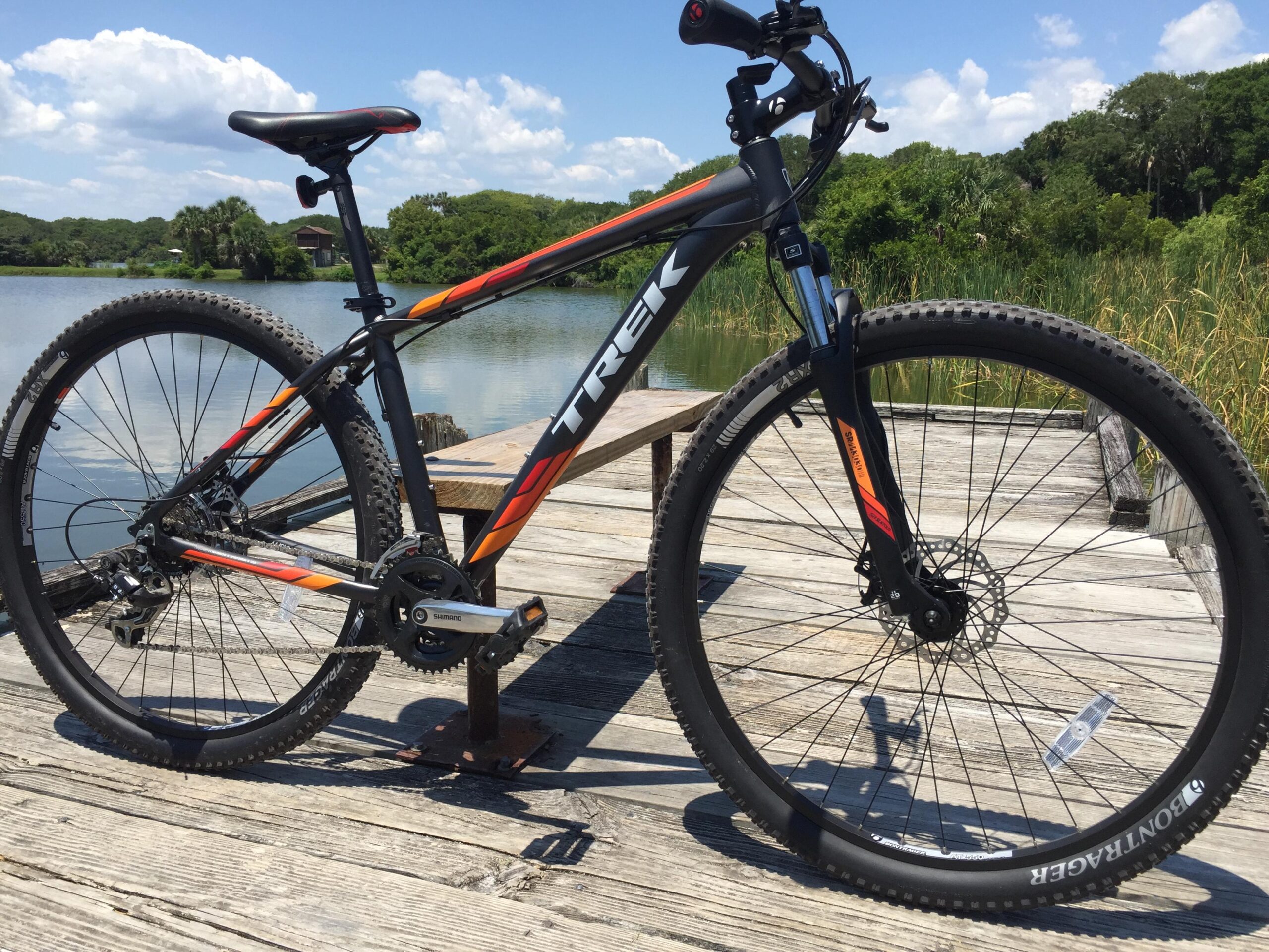 Trek Marlin 5 29er: A black and orange Trek mountain bike stands on a wooden dock by a calm lake, surrounded by greenery and under a partly cloudy sky. The bike is angled to showcase its frame, wheels, and components, including a visible chain and gears. The serene water reflects the sky, creating a peaceful outdoor scene.
