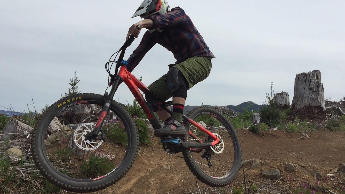 A mountain biker performs a jump on a trail, wearing a helmet and plaid shirt, with green shorts and colorful socks. The background features a wooded area with stumps and greenery, highlighting an outdoor adventurous scene. Cold Creek mountain bike trail.