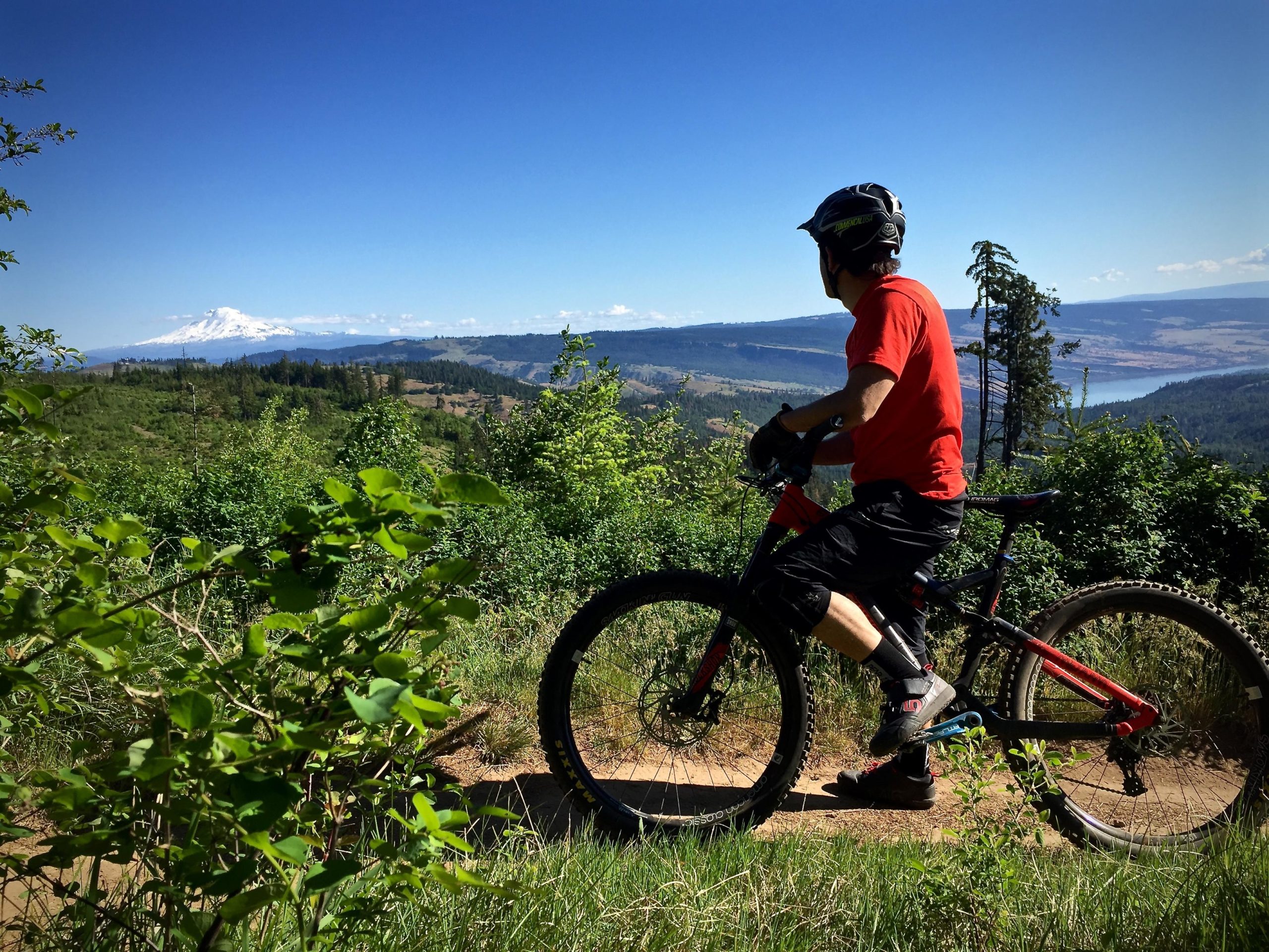 A person in a red shirt and helmet is riding a mountain bike along a trail, overlooking a scenic landscape with mountains and greenery. In the distance, a snow-capped peak is visible under a clear blue sky. Whoopdee mountain bike trail.