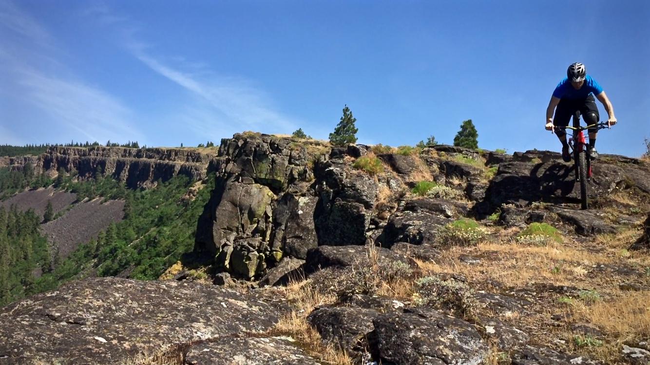 A mountain biker navigates a rocky terrain on a sunny day, with a steep drop and lush greenery in the background. The sky is clear with a few clouds, and the scene captures the excitement of outdoor adventure. Syncline mountain bike trail.