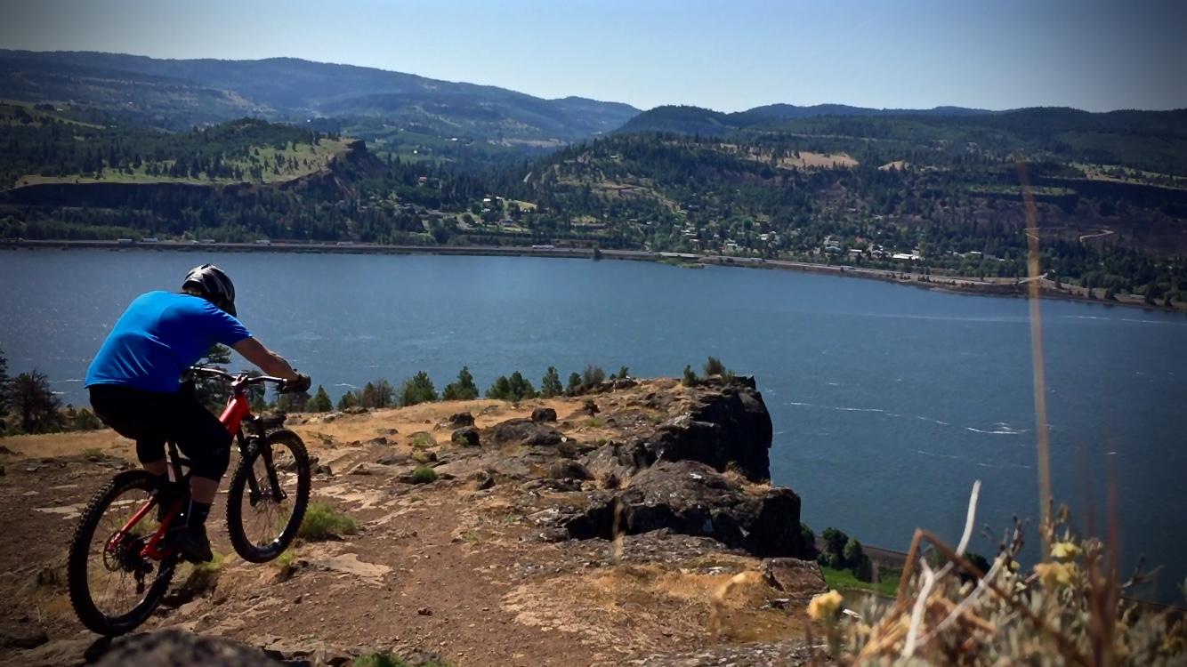 A mountain biker in a blue shirt rides down a rocky trail overlooking a scenic river and hills. The landscape features greenery and a clear blue sky. Syncline mountain bike trail.