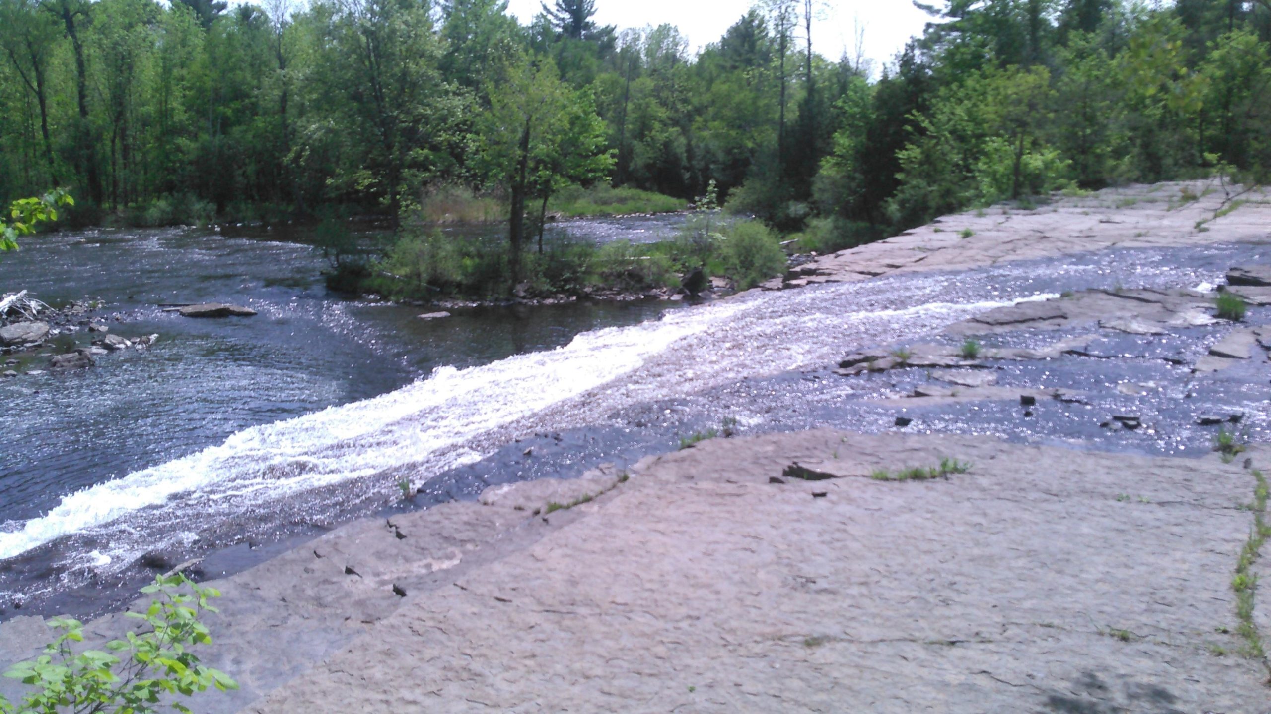 A scenic view of a gently flowing river, surrounded by lush greenery. Water cascades over a rocky ledge into the river below, with sunlight glinting off the surface. The landscape features vibrant trees and dense foliage, creating a tranquil natural setting. Blue Trail mountain bike trail.