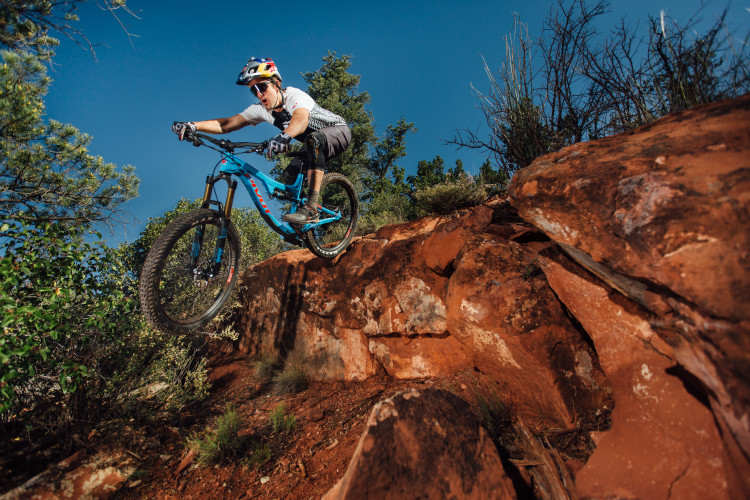 A mountain biker jumps off a rocky ledge, showcasing action and excitement against a backdrop of blue sky and green trees. The athlete wears a helmet and gear while riding a blue mountain bike, capturing the thrill of extreme sports.