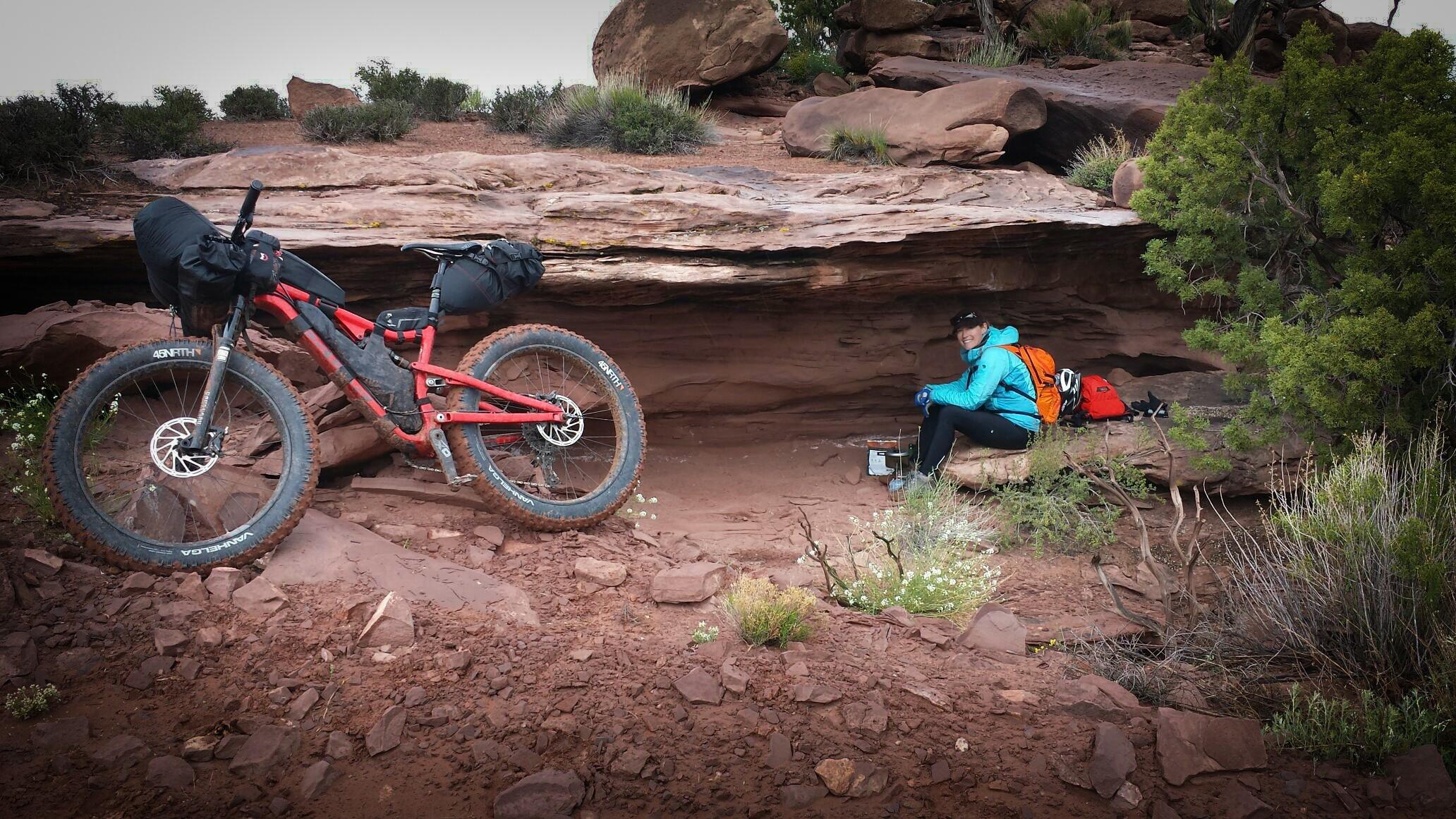 Salsa Bucksaw Carbon: A person in a blue jacket and an orange backpack sits on a rock ledge in a natural setting, preparing food with a camp stove. Nearby, a red fat bike rests on the ground, equipped for biking adventure. The scene is surrounded by rocky terrain and sparse vegetation.