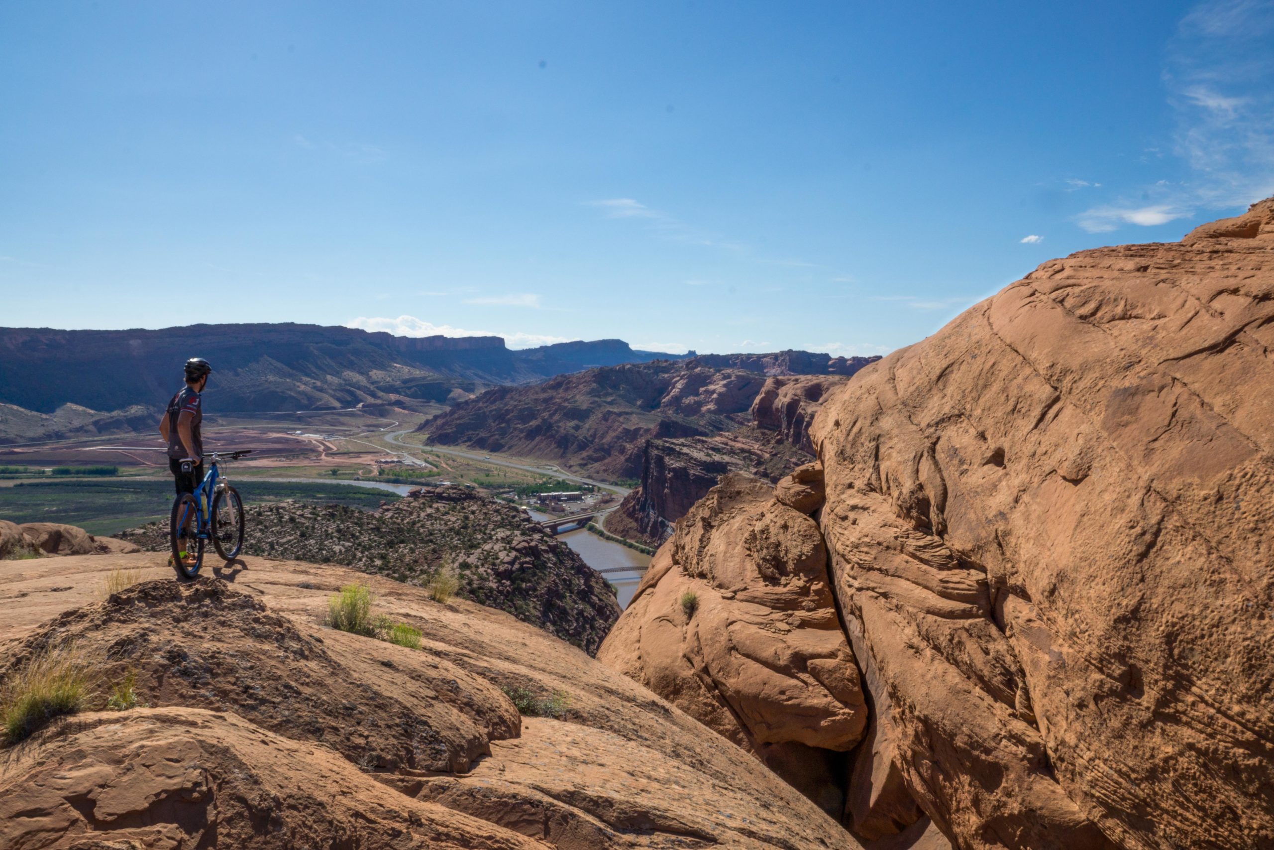A mountain biker standing on a rocky outcrop overlooking a vast landscape featuring rolling hills, a river, and a winding road in the distance under a clear blue sky. Slickrock mountain bike trail.