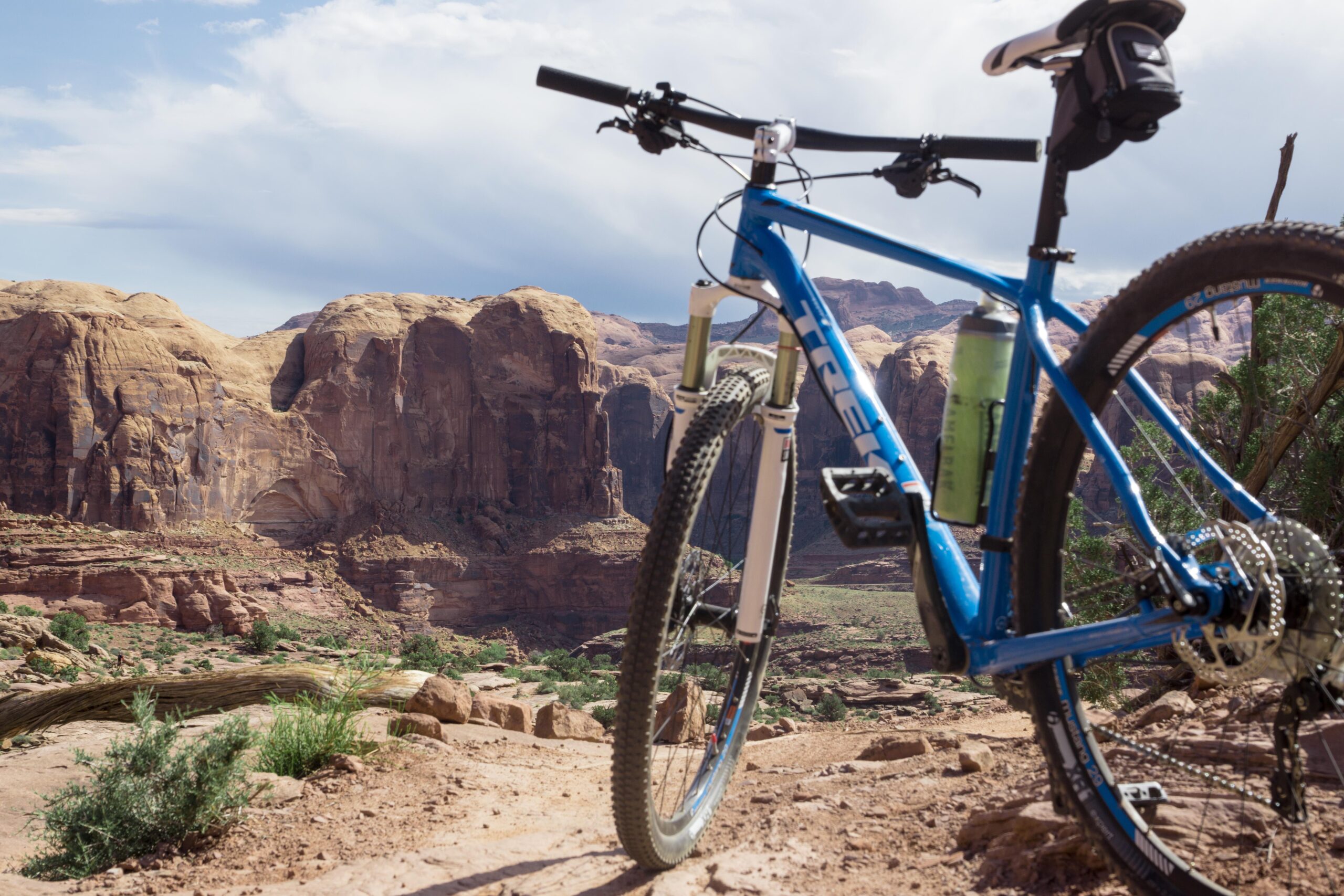 A mountain bike with a blue frame rests on a rugged dirt trail, with large rock formations and cliffs in the background. The sky above is partly cloudy, and lush greenery is visible in the surrounding landscape. Amasa Back Area mountain bike trail.