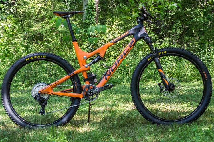 A vibrant orange and black mountain bike leaning against a lush green background, showcasing its sturdy frame, suspension system, and Maxxis tires.