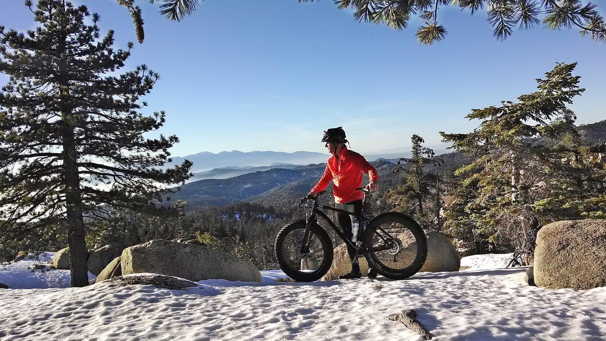 A person in a red jacket and helmet stands next to a fat-tire mountain bike on a snowy path surrounded by pine trees. In the background, sweeping mountain vistas and a clear blue sky create a scenic outdoor landscape. Exploration Trail mountain bike trail.