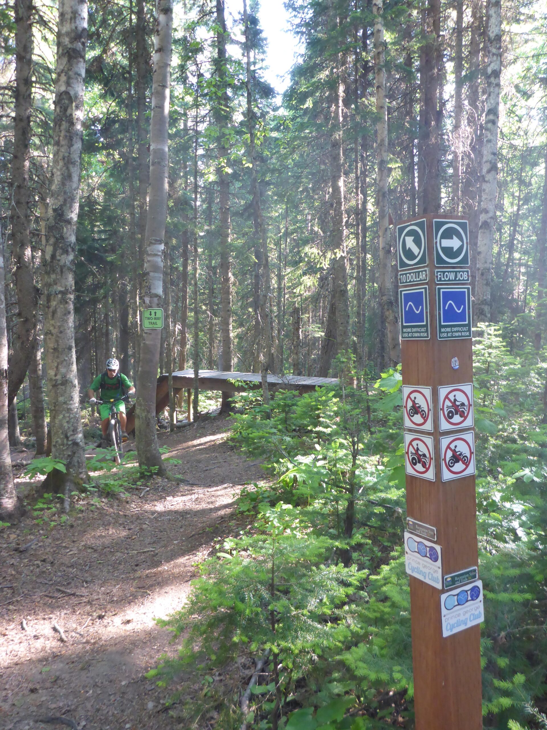 A wooded trail with a mountain biker riding in the background. In the foreground, a signpost displays trail direction signs for "10 Dollar" and "Flow Job" along with various symbols indicating trail use rules, including no motorized vehicles. Lush green foliage surrounds the dirt path, with sunlight filtering through the trees. Piderney Recreation Site mountain bike trail.