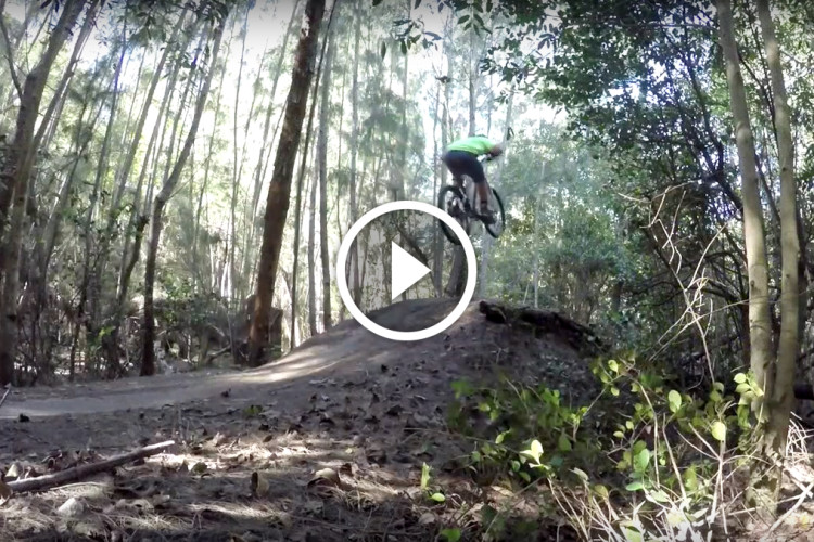 A mountain biker performing a jump over a dirt mound in a forested area, surrounded by tall trees and green foliage. A play button overlay indicates that the image is a video.