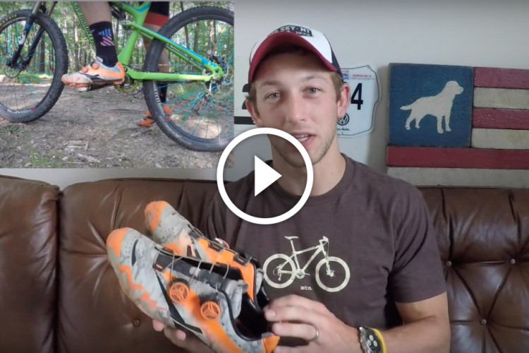 A person sitting on a brown couch holds a pair of cycling shoes, while a video preview overlay shows a mountain bike in motion on a dirt trail. The background features a wooden wall with a painted dog silhouette and a bicycle-themed shirt being worn by the individual.