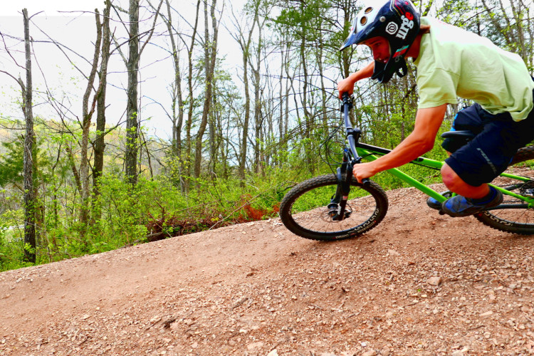 A mountain biker in a helmet leans into a turn on a dirt trail, surrounded by trees and greenery. The bike is equipped for off-road riding, and the rider is wearing casual sports attire. The setting suggests an outdoor adventure in a natural environment.