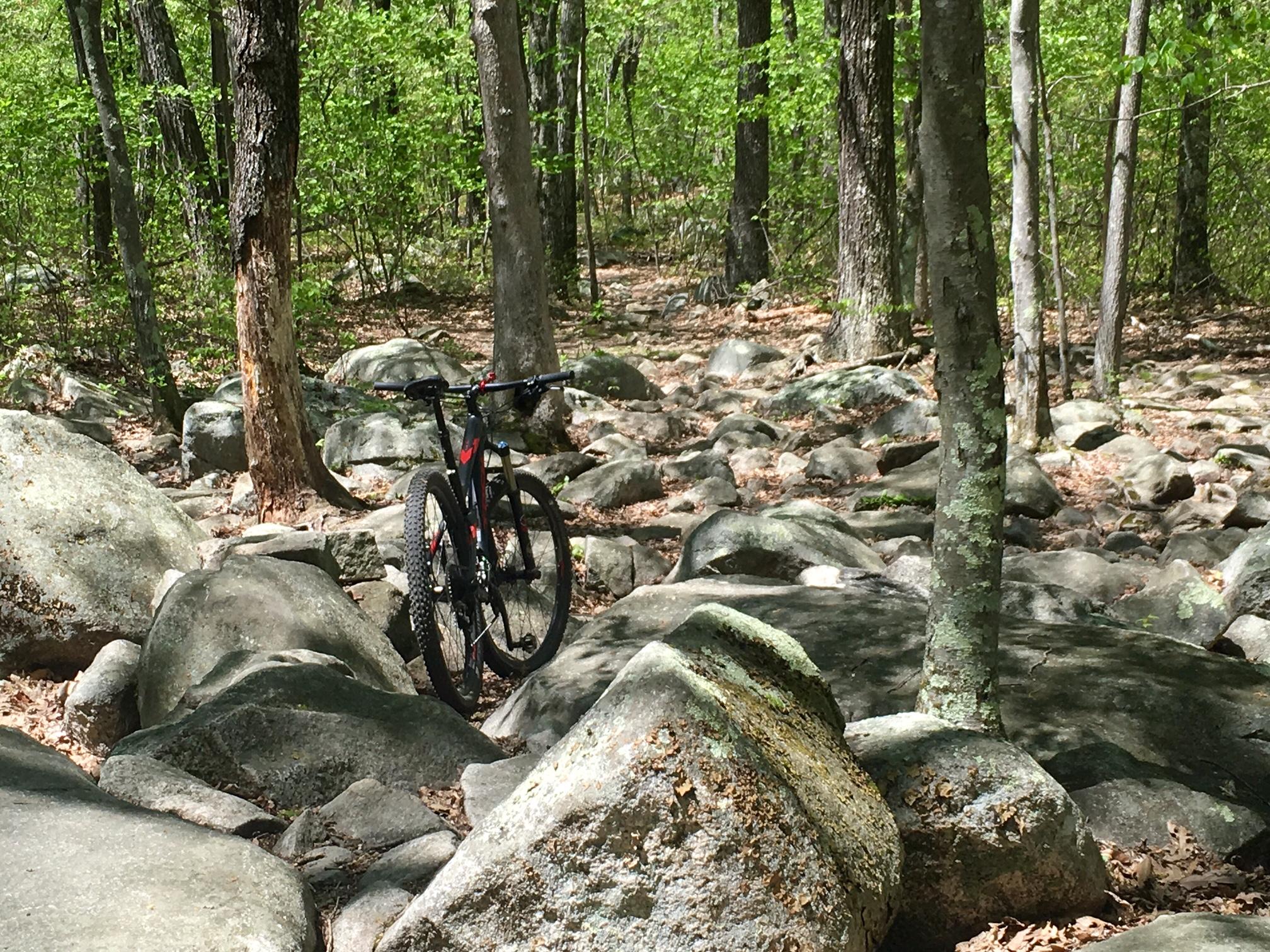A mountain bike resting on rocky terrain in a lush green forest, with trees in the background and scattered large boulders. Vietnam Trails mountain bike trail.