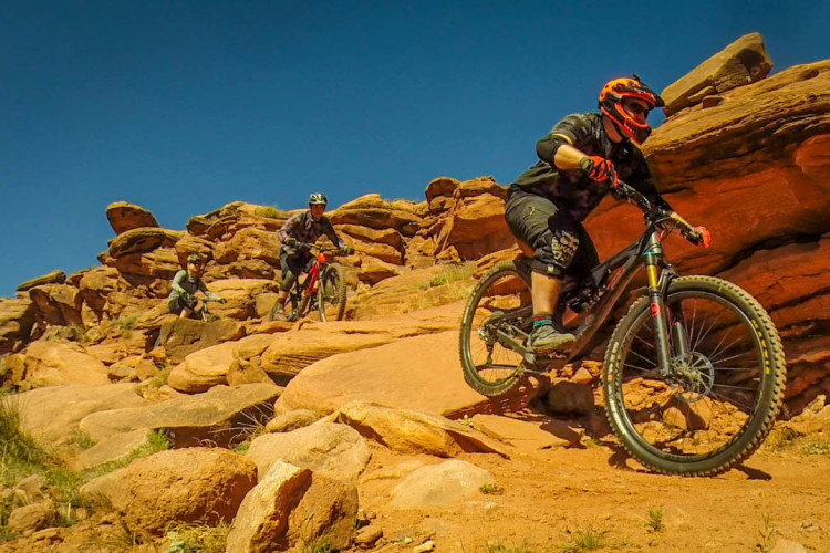 Three mountain bikers navigate a rocky, desert terrain under a clear blue sky. The cyclist in the foreground rides downhill, expertly maneuvering around the boulders, while two others are positioned further back, one pausing to assess the path. The landscape features red rock formations, with patches of grass visible among the stones.