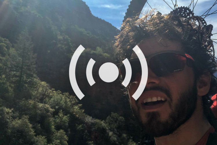 A person with curly hair and sunglasses smiles for a selfie, surrounded by lush green mountains and bright sunlight.
