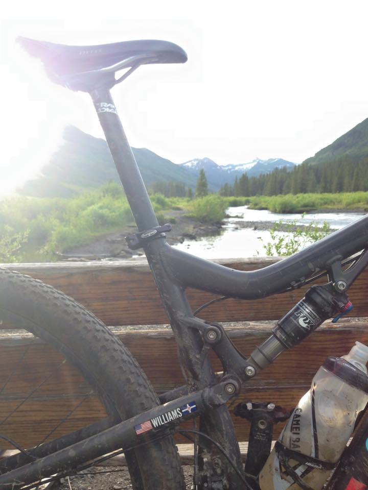Santa Cruz Tallboy: A close-up view of a mountain bike leaning against a wooden railing, with a scenic landscape in the background. The image captures the bike's dirty frame and tire, with a visible water bottle attached. In the distance, lush green hills and snow-capped mountains can be seen under a bright sky.