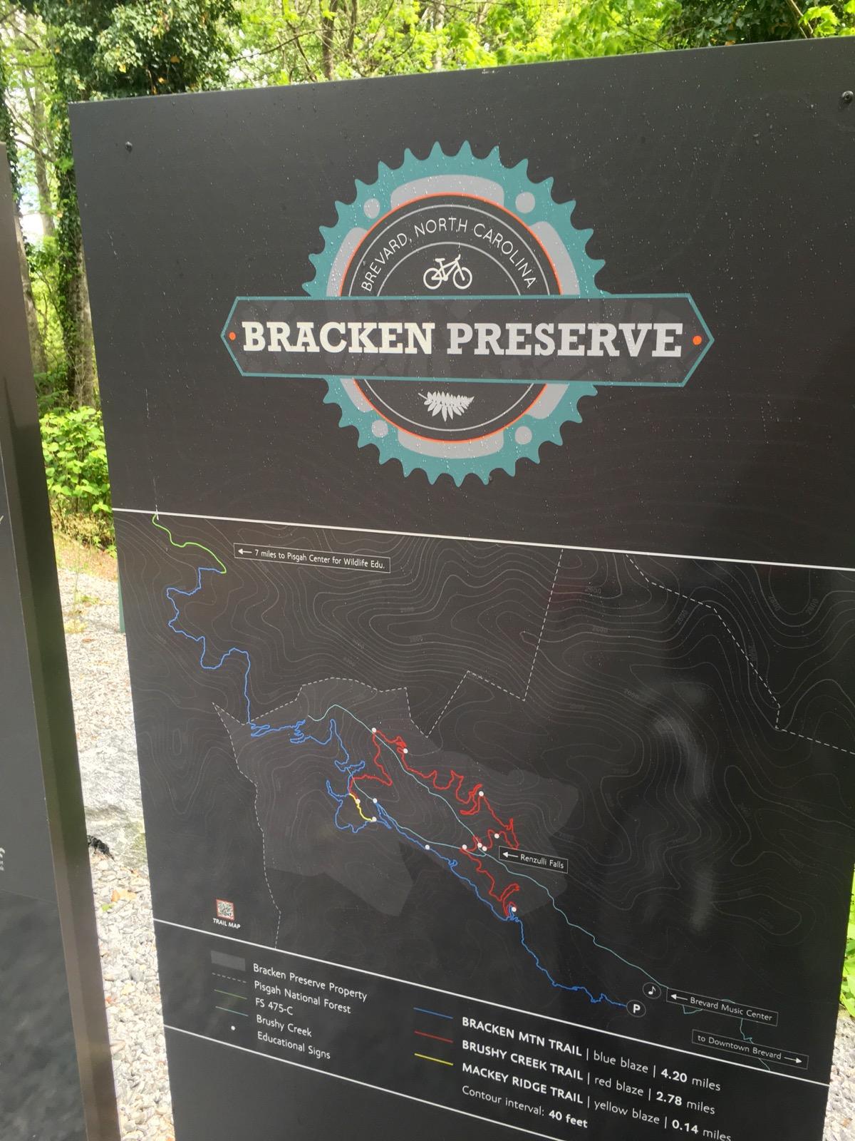 Map and information sign for Bracken Preserve in Brevard, North Carolina, featuring trail details including the Bracken Mountain Trail, Brushy Creek Trail, and Mackey Ridge Trail. The sign includes a visual representation of the trails marked in blue, red, and yellow, along with contour information and directions to nearby landmarks. Bracken Preserve mountain bike trail.