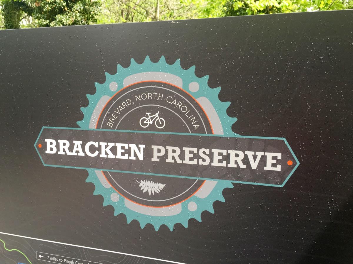 Sign for Bracken Preserve in Brevard, North Carolina, featuring a decorative gear design and a bicycle icon. The sign includes the words "Bracken Preserve" prominently displayed, along with information about the location. There are water droplets on the surface, indicating recent rain. Bracken Preserve mountain bike trail.
