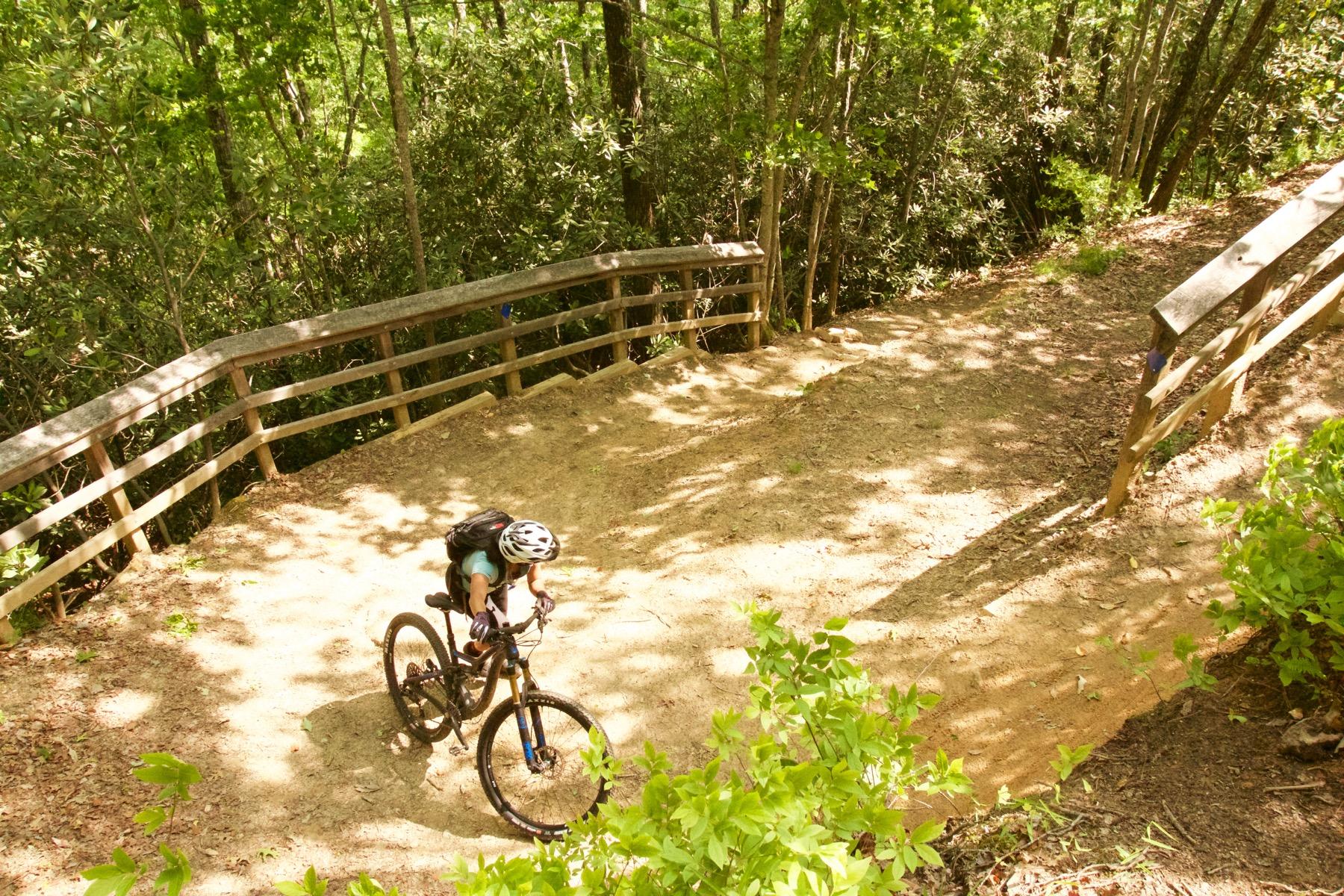 A mountain biker navigating a dirt trail surrounded by lush greenery, with a wooden railing in the background. The rider is focused on the path ahead, showcasing an adventurous outdoor scene in a natural setting. Bracken Preserve mountain bike trail.