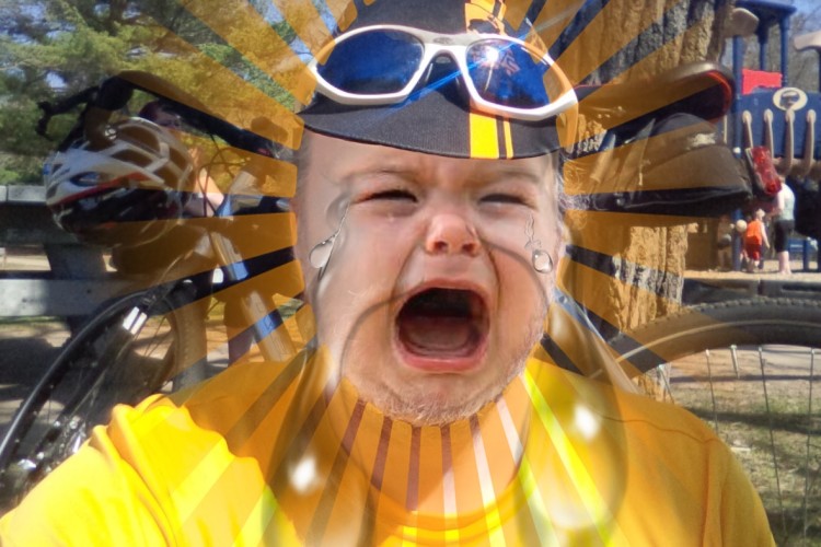 A digitally altered image of a person wearing sunglasses and a yellow shirt, with a crying facial expression overlaid and a vibrant sunburst effect in the background. Bicycles and playground equipment are visible behind them, suggesting an outdoor setting.