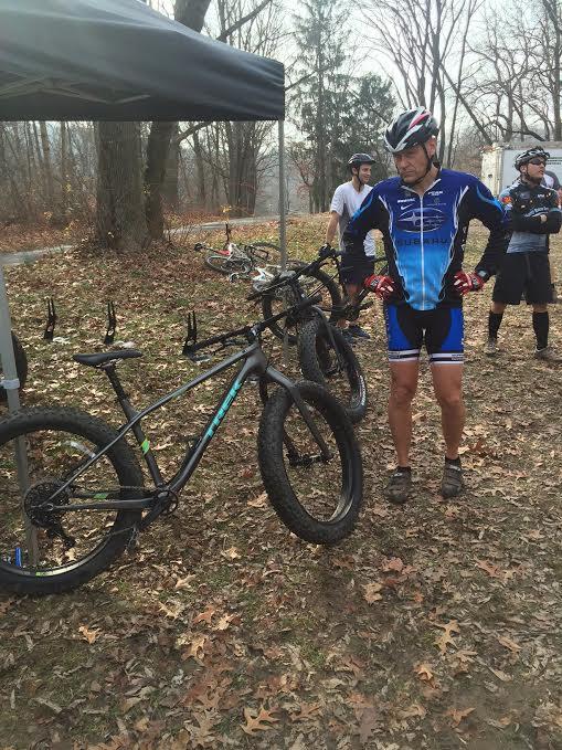 A group of mountain bike riders preparing for a ride in a wooded area, with autumn leaves scattered on the ground. One rider in a blue and black cycling outfit stands next to a fat bike, while two other riders are in the background by a tent. The scene is set in a cloudy, overcast environment.