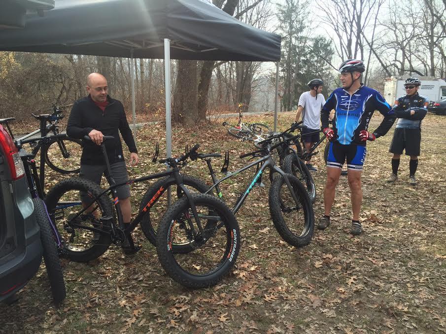 Group of cyclists gathered around fat bikes under a canopy in a wooded area. One cyclist is inspecting a bike while others are engaged in conversation, with fallen leaves covering the ground.