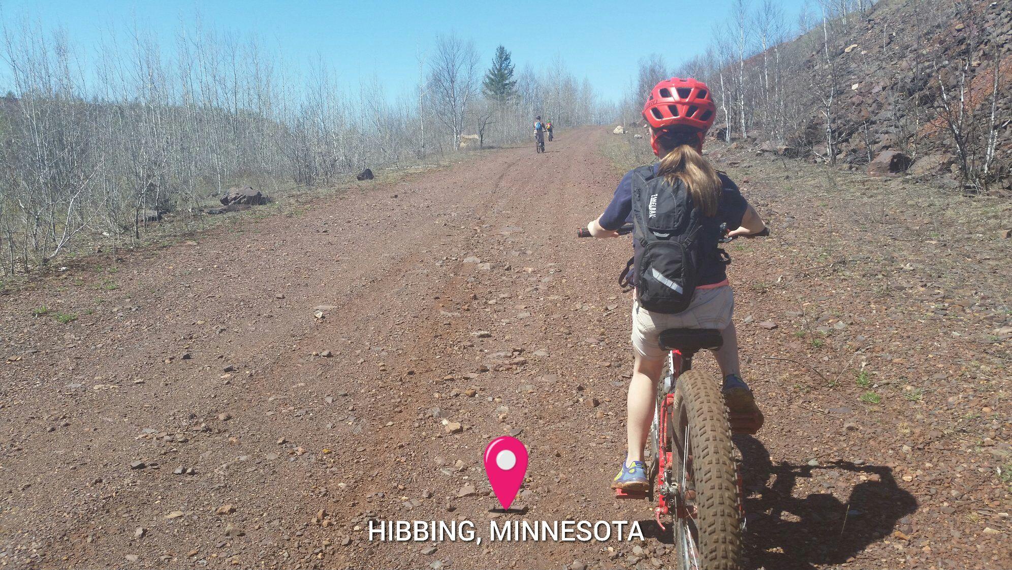 A young cyclist riding a fat bike on a gravel trail in Hibbing, Minnesota. The scene features sparse trees lining the path and another cyclist in the distance, with a clear blue sky overhead. A location marker indicates the area as Hibbing, Minnesota.