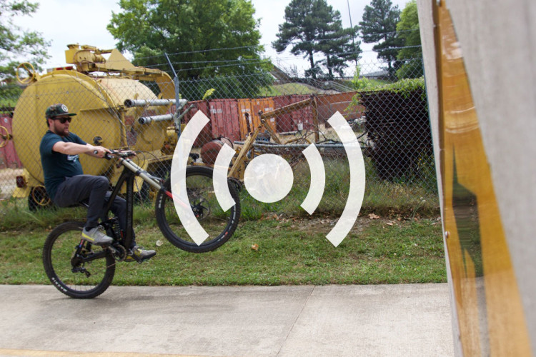 A person riding a mountain bike is performing a wheelie on a paved surface, with a chain-link fence and industrial structures in the background. The image includes graphic symbols suggesting sound or transmission.