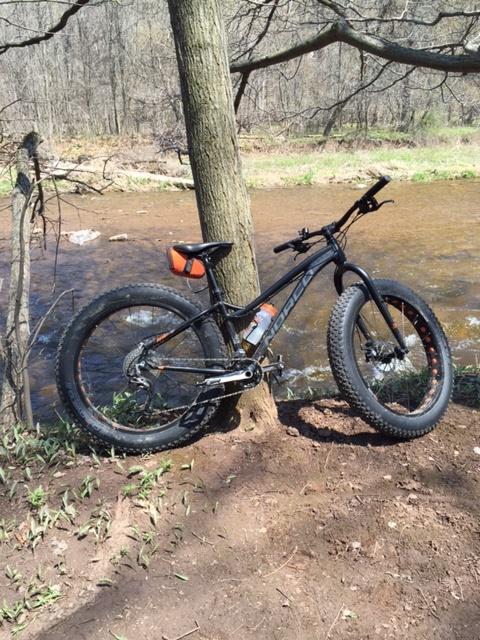 Norco Bigfoot: A black mountain bike with thick tires leans against a tree by a small river in a wooded area, with spring greenery visible on the ground. The scene captures a moment of outdoor adventure amidst nature.