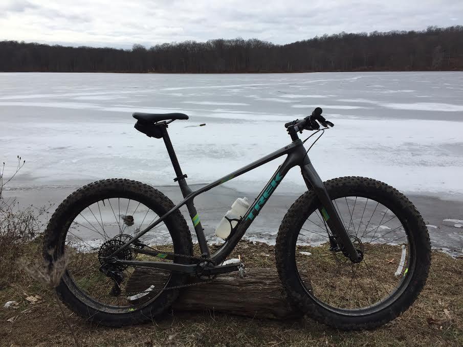 A fat tire mountain bike is parked on a log near a frozen lake, with a cloudy sky above and trees in the background.