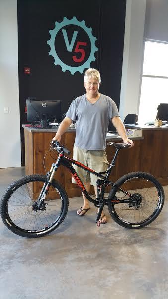 A man standing next to a mountain bike inside a shop, wearing a gray t-shirt and shorts. The background features a logo that says "V5" and a counter with a computer. The bike is black with red accents and has thick tires suitable for off-road riding.