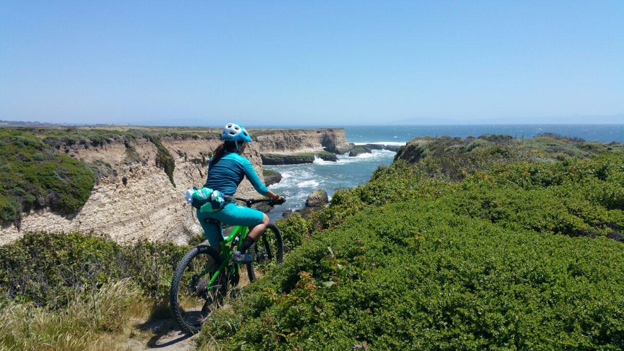 A person in a blue and green cycling outfit is riding a mountain bike along a coastal trail, overlooking rugged cliffs and the ocean. The sky is clear and sunny, with gentle waves crashing against the rocks below. Lush green vegetation surrounds the pathway, creating a scenic backdrop. Wilder Ranch State Park mountain bike trail.