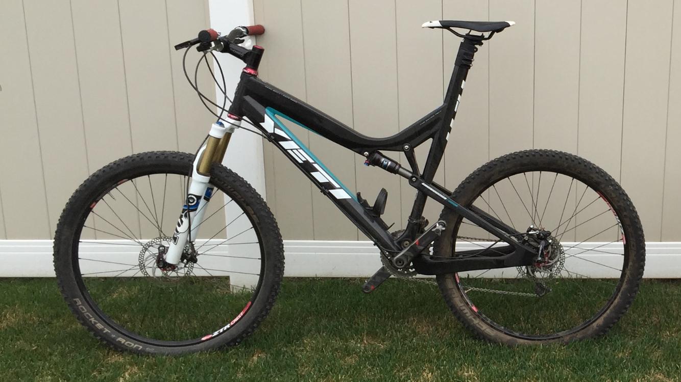 Yeti ASRc: A black and blue mountain bike with visible suspension components, standing on grass in front of a light-colored wall. The bike features thick, knobby tires and a sleek design typical of high-performance mountain bikes.
