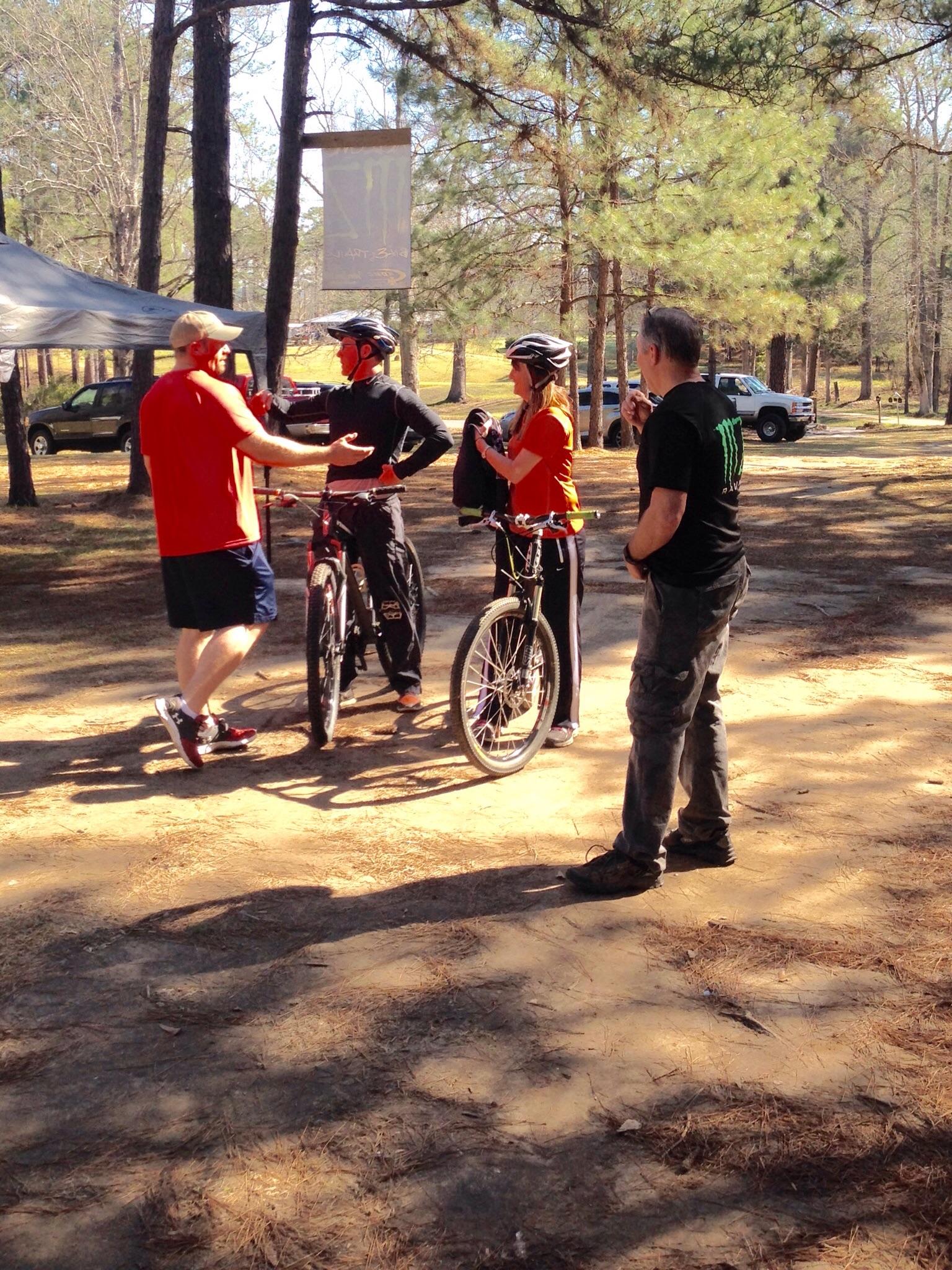 A group of four people, two wearing biking helmets, stand near bicycles in a wooded area. They are engaged in conversation, with one person gesturing. The setting features tall pine trees and a tent in the background, suggesting a casual outdoor event. A car is parked in the distance, and a Monster Energy banner hangs above. The group appears to be enjoying a sunny day outdoors. Mt. Zion Bike Trails mountain bike trail.