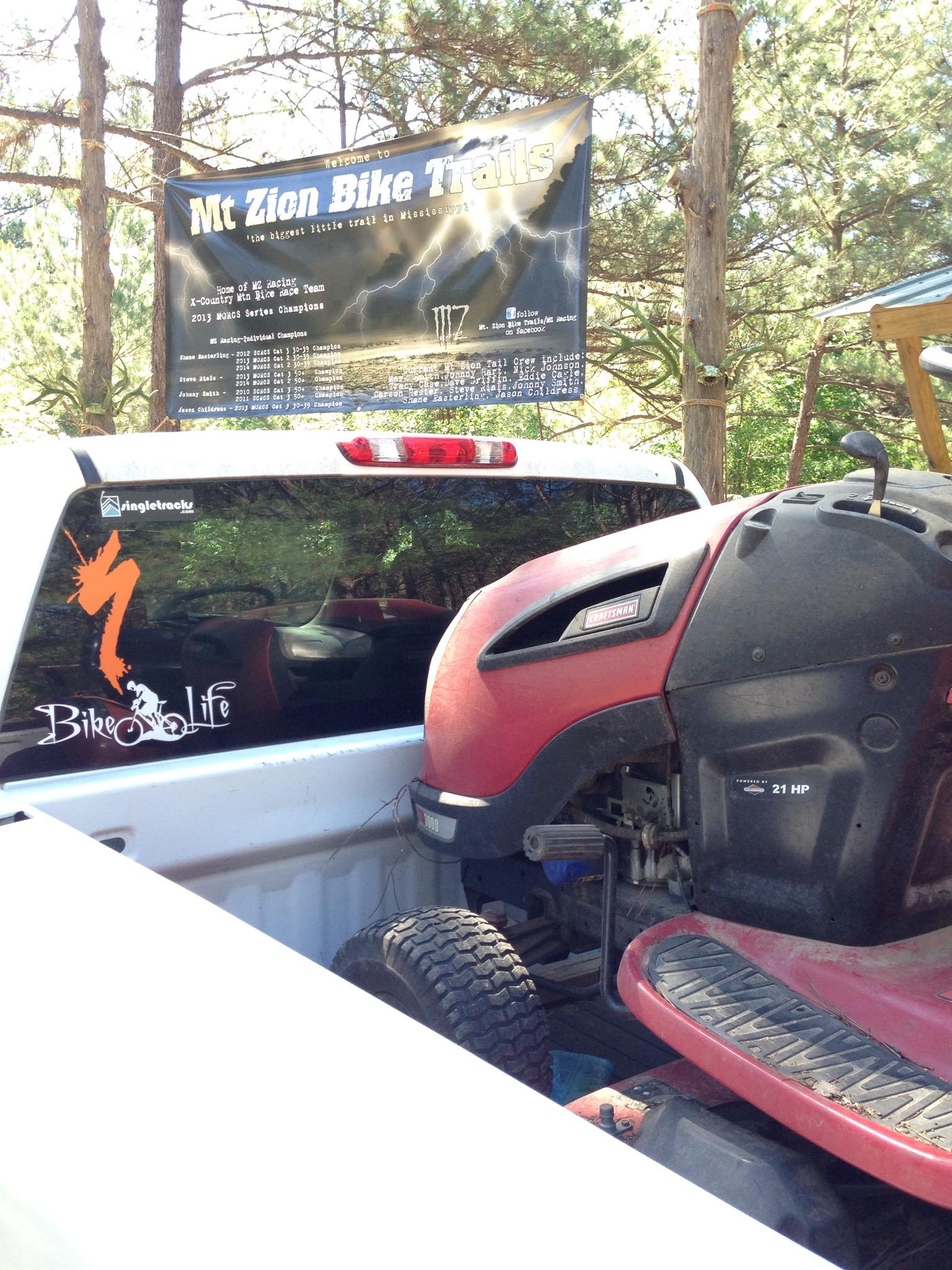 A pickup truck with a visible banner reading "Welcome to Mt Zion Bike Trails," set among trees. The truck bed contains a red lawn mower with a visible engine compartment. A sticker on the rear window features the phrase "Bike Life." Mt. Zion Bike Trails mountain bike trail.