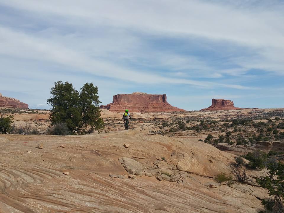 A biker wearing a bright helmet rides along a rocky terrain with desert vegetation, set against a backdrop of large mesas and a clear blue sky. Ramblin' mountain bike trail.