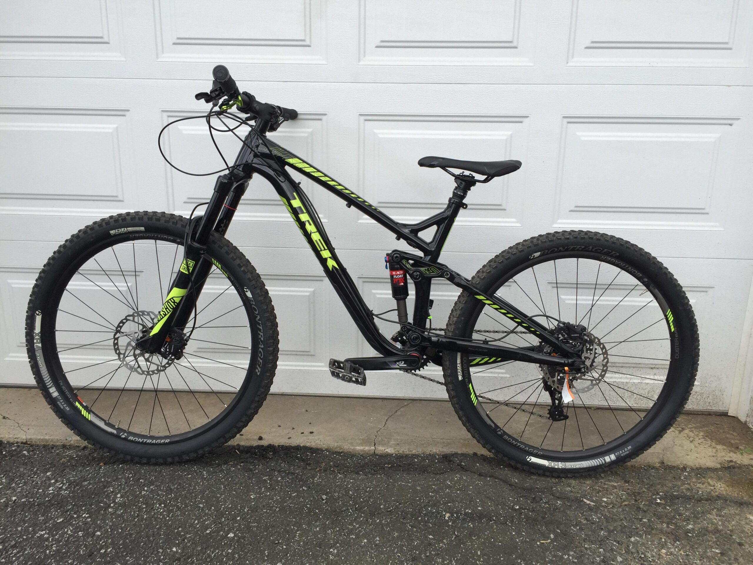 Trek Remedy 8 27.5/650b: A black mountain bike with bright green accents, featuring a full suspension design and large knobby tires, positioned against a white garage door. The bike has disc brakes, a sporty frame, and is equipped for off-road riding.